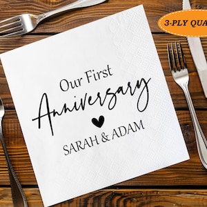 Può includere: Un tovagliolo di carta bianco con la scritta "Our First Anniversary" in corsivo nero, un cuore nero e i nomi "SARAH & ADAM". Posate sono disposte attorno al tovagliolo su un tavolo di legno.