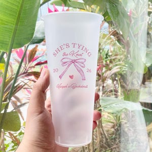 以下が含まれることがあります： ピンクのグラデーション効果のある、つや消しプラスチックカップ。カップには、リボン模様と数字「20」と「26」とともに「SHE'S TYING the Knot」の文字がプリントされています。テキストの下には「Harper's Bachelorette」と書かれています。