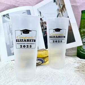 May include: Two white frosted plastic cups with black and gold graduation themed designs. The text on the cups reads "Congrats! Elizabeth Princeton University 2025".