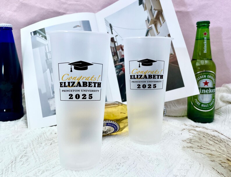 May include: Two white frosted plastic cups with black and gold graduation themed designs. The text on the cups reads "Congrats! Elizabeth Princeton University 2025".