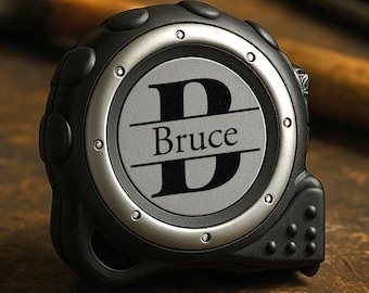 May include: A black and silver tape measure with the name "Bruce" and the letter "B" engraved on the face. A hammer and other tools are in the background, suggesting a construction or DIY theme.