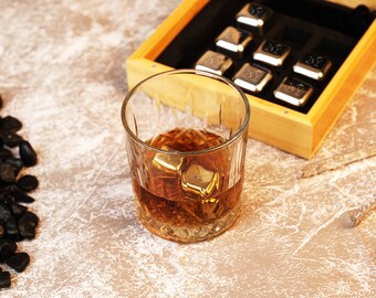 May include: A glass of whiskey with ice cubes in a clear glass on a light brown surface. A wooden box with metal ice cubes is next to the glass.