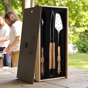 May include: A wooden box set containing grilling tools. The set includes a spatula, knife, and tongs with wooden handles. The box is engraved with the text "Ferdinand Grill Master". The tools are displayed against a black background.