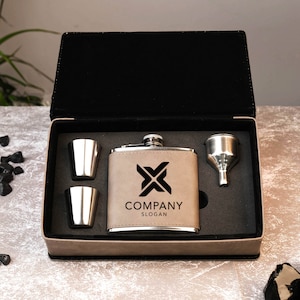 May include: A brown leather flask with a silver top and a black logo that says "COMPANY SLOGAN". The flask is in a black box with two silver shot glasses and a silver funnel.