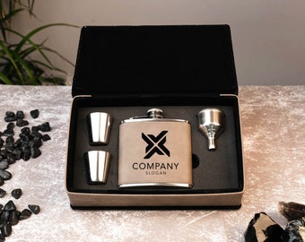 May include: A brown leather flask with a silver top and a black logo that says "COMPANY SLOGAN". The flask is in a black box with two silver shot glasses and a silver funnel.