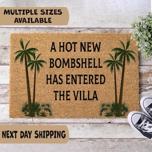 May include: A rectangular tan coir doormat featuring black text that reads A HOT NEW BOMBSHELL HAS ENTERED THE VILLA. The design includes two green palm trees on the left and two green palm trees on the right side of the text, centered on the mat.