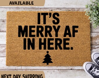 May include: A rectangular doormat made of natural coir fiber. The mat features the phrase "IT'S MERRY AF IN HERE" in bold black letters, with a black Christmas tree graphic below. The words "MULTIPLE SIZES AVAILABLE" are at the top.