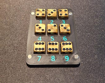 May include: A black game board with nine gold-coloured dice, each displaying a different number of black dots. The numbers 1 to 9 are printed in teal on the board. The dice are arranged in a 3x3 grid.