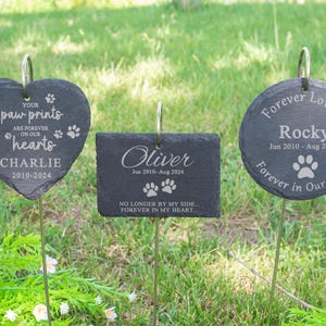Puede incluir: Tres marcadores conmemorativos para mascotas de pizarra gris oscuro en forma de corazón, rectangular y circular. Cada uno tiene una estaca de metal y presenta texto grabado y huellas de patas. El marcador en forma de corazón dice "Tus huellas de patas están para siempre en nuestros corazones, Charlie 2010-2024."