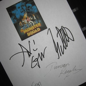 Peut inclure: Une feuille de papier blanche avec une image en noir et blanc d'une affiche de film pour "The Monster Squad" avec le texte "The Monster Squad" en jaune et rouge. Le papier est signé par trois personnes : "A.L. Ewing", "Duncan Regehr" et "Tom", la dernière signature étant en plus petit.