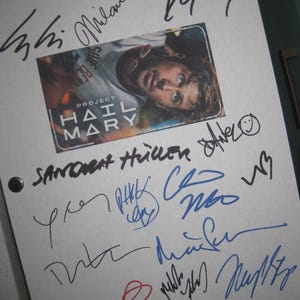 May include: A white sheet of paper covered in signatures in black, blue, and red ink. A small image from "Project Hail Mary" is in the centre. The image shows a man with a surprised expression. The paper is held together with two black binder rings.