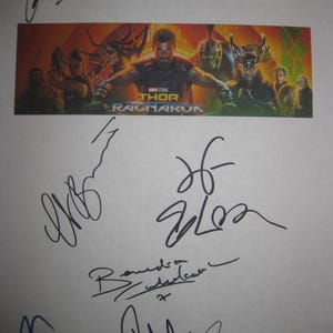 Può includere: Un foglio bianco con più firme a inchiostro nero. Una striscia colorata presenta il poster del film "Thor: Ragnarok". Le firme sono di vari stili e dimensioni, alcune sovrapposte.