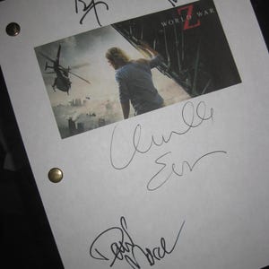 May include: A white paper with a printed image from the movie "World War Z" featuring a person and helicopters. The paper is signed by multiple people with black ink. Gold-colored fasteners secure the paper.