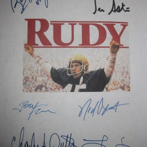 May include: A print featuring the word "RUDY" in red letters above an image of a football player in a helmet. The print is signed by multiple people in blue ink.