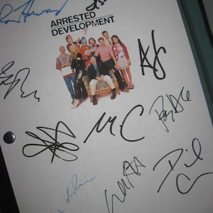 Puede incluir: Un guion blanco con el título "Arrested Development" y una foto grupal del elenco. El guion está cubierto con múltiples firmas en tinta negra y azul.