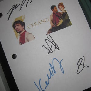 May include: A white paper with the word "CYRANO" in gold lettering, featuring images of actors and multiple signatures in black and blue ink. The paper is held in a black binder.