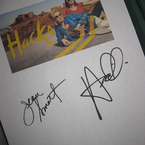 Può includere: Un foglio di carta bianco con due firme, una in inchiostro nero che dice "Jean Smart" e l'altra in inchiostro nero che dice "Hacks". Il foglio ha anche una foto di due donne in un ambiente desertico con la parola "Hacks" in lettere gialle.