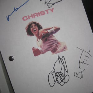 May include: A white paper with the title "CHRISTY" in pink letters, featuring a photo of a woman in boxing gloves. The paper is signed by multiple people in black and blue ink. Two black rivets are visible on the left side.