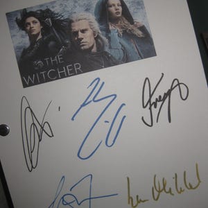 May include: A white sheet of paper with a printed image from "The Witcher" series, featuring three characters and the show's title. Several signatures in black, blue, and gold ink are present on the paper, likely autographs.