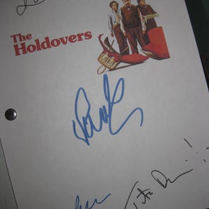 May include: A white sheet of paper featuring the movie title "The Holdovers" in red letters, accompanied by a movie poster illustration and multiple signatures in blue and black ink. The paper has two black metal grommets.