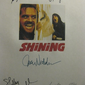 Puede incluir: Un cartel de la película "El Resplandor" con una imagen central de Jack Nicholson y Shelley Duvall. El título "SHINING" está en rojo y hay varias firmas presentes.