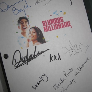 Op de afbeelding: Een wit vel papier met meerdere handtekeningen van de cast van de film "Slumdog Millionaire". De handtekeningen omvatten Dev Patel, Frieda Pinto en de regisseur Danny Boyle. De titel van de film is in rood en wit op het papier gedrukt.