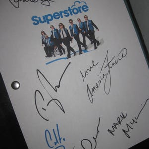 May include: A white paper with the word "Superstore" in blue, along with a photo of a group of people in suits and sunglasses. The paper is covered in signatures in black and blue ink.