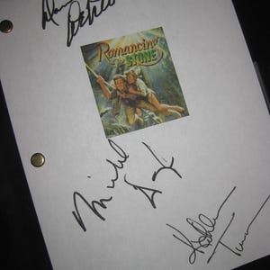 Może przedstawiać: Biała kartka papieru z plakatem filmu "Miłość, szmaragd i krokodyl". Plakat przedstawia mężczyznę i kobietę w scenie przygody. Papier jest podpisany przez kilka osób czarnym tuszem.