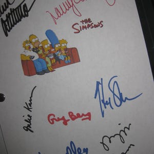 Puede incluir: Una hoja de papel blanca con una imagen de dibujos animados de la familia Simpson en un sofá rojo. La hoja está cubierta de firmas en tinta negra, azul y roja, incluyendo los nombres "Julie Kavner" y "Yeardley Smith".