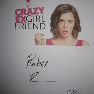 May include: A white poster with the title "Crazy Ex-Girlfriend" in black text. The poster features a woman with brown hair and a pink shirt holding a pink balloon. The text "Never. Let. Go." is at the top of the poster. The poster is signed "Rachel" and "Sada" in black ink.