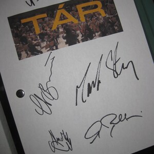 May include: A white sheet of paper with the word "TÁR" in yellow letters over an image of an orchestra. The paper is signed in black ink by multiple people. The paper is held in a black binder.