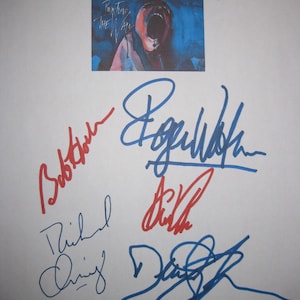 Puede incluir: Papel blanco con una pequeña imagen de la portada del álbum con el texto "Pink Floyd The Wall". La imagen está rodeada de múltiples firmas en tinta azul y roja.