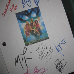 May include: A white document featuring a central Scooby-Doo movie poster with the cast and a shadowy figure. The document is surrounded by multiple handwritten signatures in blue, black, and red ink, including the text "Ruh-roh! Voice of Scooby-Doo!"