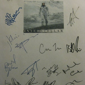 May include: A white sheet of paper with black ink signatures from the cast and crew of the movie Interstellar. The signatures include: Ellen Burstyn, Matt Damon, Jessica Chastain, Anne Hathaway, David Gyllenhaal,  and others. The movie poster for Interstellar is in the background.