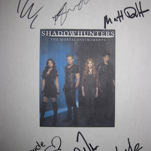 Könnte beinhalten: Ein gedrucktes Bild von "Shadowhunters: The Mortal Instruments" mit den Darstellern. Das Bild ist von handschriftlichen Unterschriften in schwarzer Tinte umgeben. Der Titel der Show ist in weißen Buchstaben auf dunklem Hintergrund gedruckt.