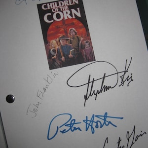 Puede incluir: Una hoja de papel blanca con un cartel de la película "Children of the Corn" y múltiples firmas en tinta azul y negra. El cartel muestra a un grupo de personas en un campo con un cielo rojo.