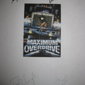 May include: A movie poster for "Maximum Overdrive" featuring a menacing semi-truck with the film's title in bold, blue and white lettering. The poster is signed by multiple people in black ink.