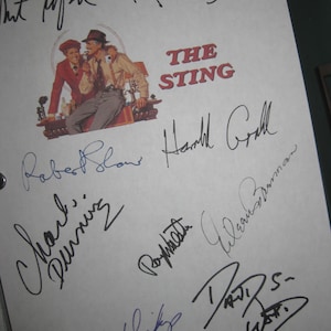 Op de afbeelding: Een wit vel met talrijke handtekeningen in zwarte en blauwe inkt. De titel "THE STING" is in het rood gedrukt, vergezeld van een illustratie van twee mannen. De handtekeningen zijn van de cast en crew van de film.
