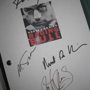 Op de afbeelding: Een wit vel papier met de filmposter voor "Raging Bull" met Robert De Niro. De poster is in zwart, wit en rood. Het papier heeft meerdere handtekeningen in zwarte inkt.