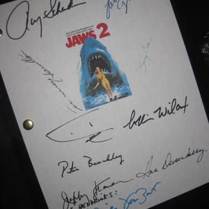 Puede incluir: Un papel blanco con múltiples firmas, incluyendo "Amy Scheid" y "Lorraine Gary". El papel presenta una imagen del póster de la película "JAWS 2" de un tiburón con una mujer en el agua. También es visible el texto "Just when you thought it was safe to go back in the water...".