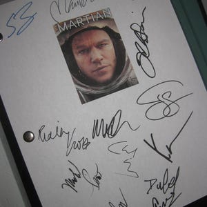 May include: A white sheet with multiple signatures in black ink. A small image from the movie "The Martian" is in the center. The image features Matt Damon in a space suit. The sheet is held together with two silver rivets.