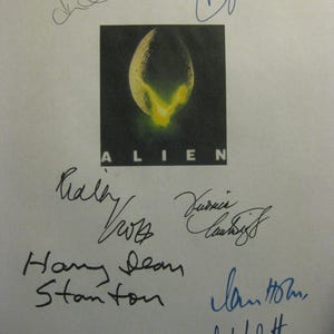 May include: A white sheet of paper featuring the movie title "Alien" with the iconic egg image. The paper is covered with multiple signatures in black and blue ink, likely autographs from the film's cast and crew.
