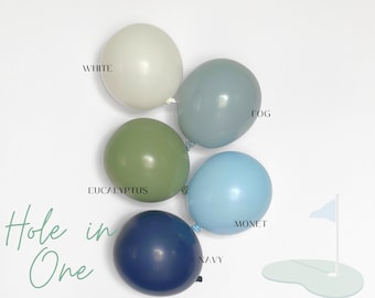 May include: Five balloons in white, fog, eucalyptus, monet, and navy colors. The text BLOOM LUXURY BALLOONS appears at the top, and the text Hole in One is written in script on the bottom left. A small graphic of a golf flag and hole is visible.