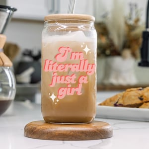 May include: A glass jar with a wooden lid filled with iced coffee. The jar has a pink and white sticker that says "I'm literally just a girl".