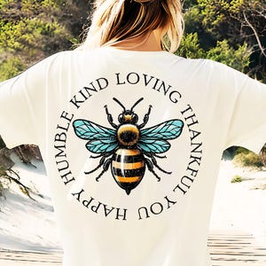 Puede incluir: Camiseta blanca con un gráfico circular negro que presenta una abeja con rayas amarillas y negras y alas azules. El círculo también tiene el texto "Happy Humble Kind Loving Thankful You" escrito alrededor de la abeja.