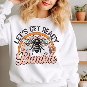 Könnte beinhalten: Weißes Sweatshirt mit einer Grafik mit einer Biene, einem Regenbogen und dem Text "LET'S GET READY TO BUMBLE" in Schwarz, Orange und Rosa. Das Design ist auf der Vorderseite des langärmeligen Oberteils zentriert.