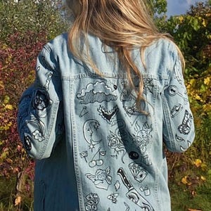 May include: A light blue denim jacket featuring a variety of black and white hand-drawn illustrations on the back and sleeves. The designs include a crescent moon, a knife, a skull, roses, cherries, dice, playing cards, and various small symbols.