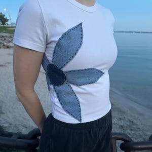 Peut inclure: Un t-shirt blanc à manches courtes orné d'une grande fleur appliquée en tissu denim bleu clair. Les pétales et le centre de la fleur sont fixés sur le côté du t-shirt avec des coutures apparentes en fil foncé le long des bords.