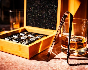 May include: A wooden box with a set of silver ice cubes and a glass of amber liquid with ice cubes and a silver ice cube tongs.