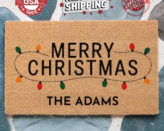 May include: A rectangular doormat made of natural coir fiber. The mat features the words "MERRY CHRISTMAS" in large black letters, with a string of colorful Christmas lights above the text. The family name "THE ADAMS" is printed below.